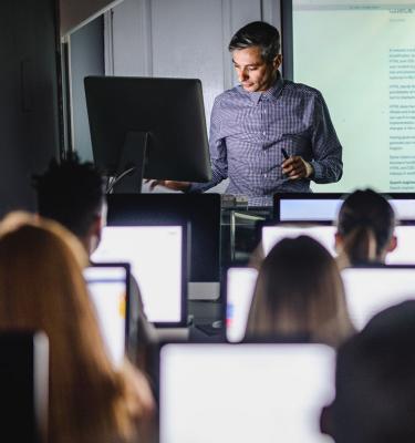 A man stands at the front of a classroom, presenting something on a screen.